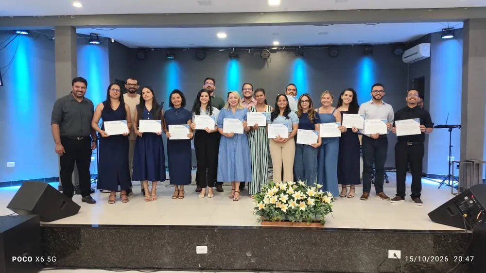 Formandos da turma 2026.1 enfileirados no palco, segurando certificados, com um arranjo de flores brancas à frente