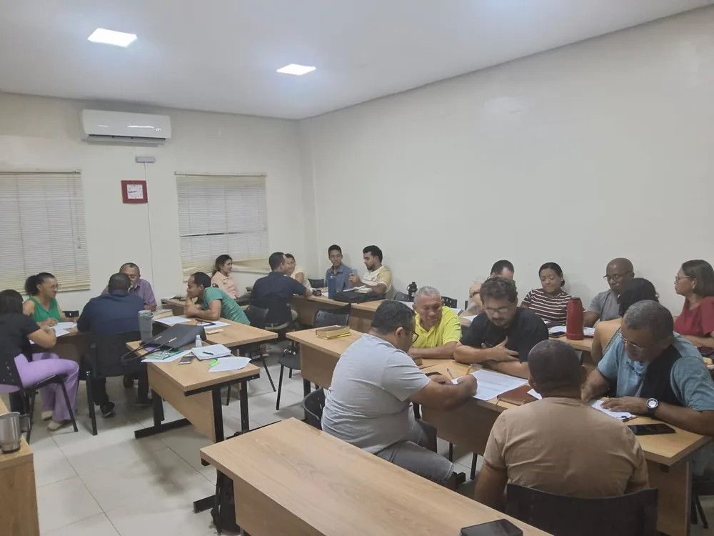 Alunos adultos reunidos em mesas, em grupos, conversando e lendo folhas durante uma atividade em sala de aula