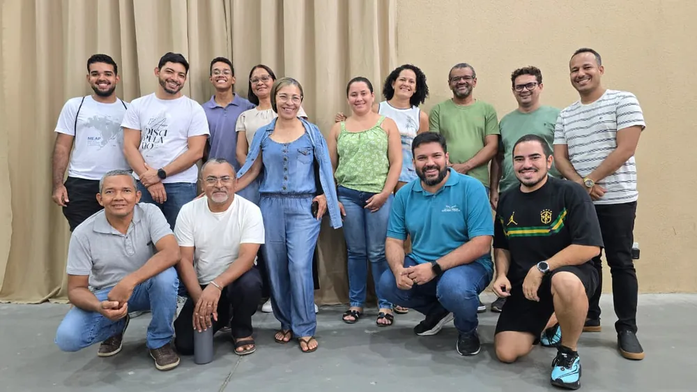 Um grupo de adultos sorrindo, em pé e agachados, diante de uma cortina bege